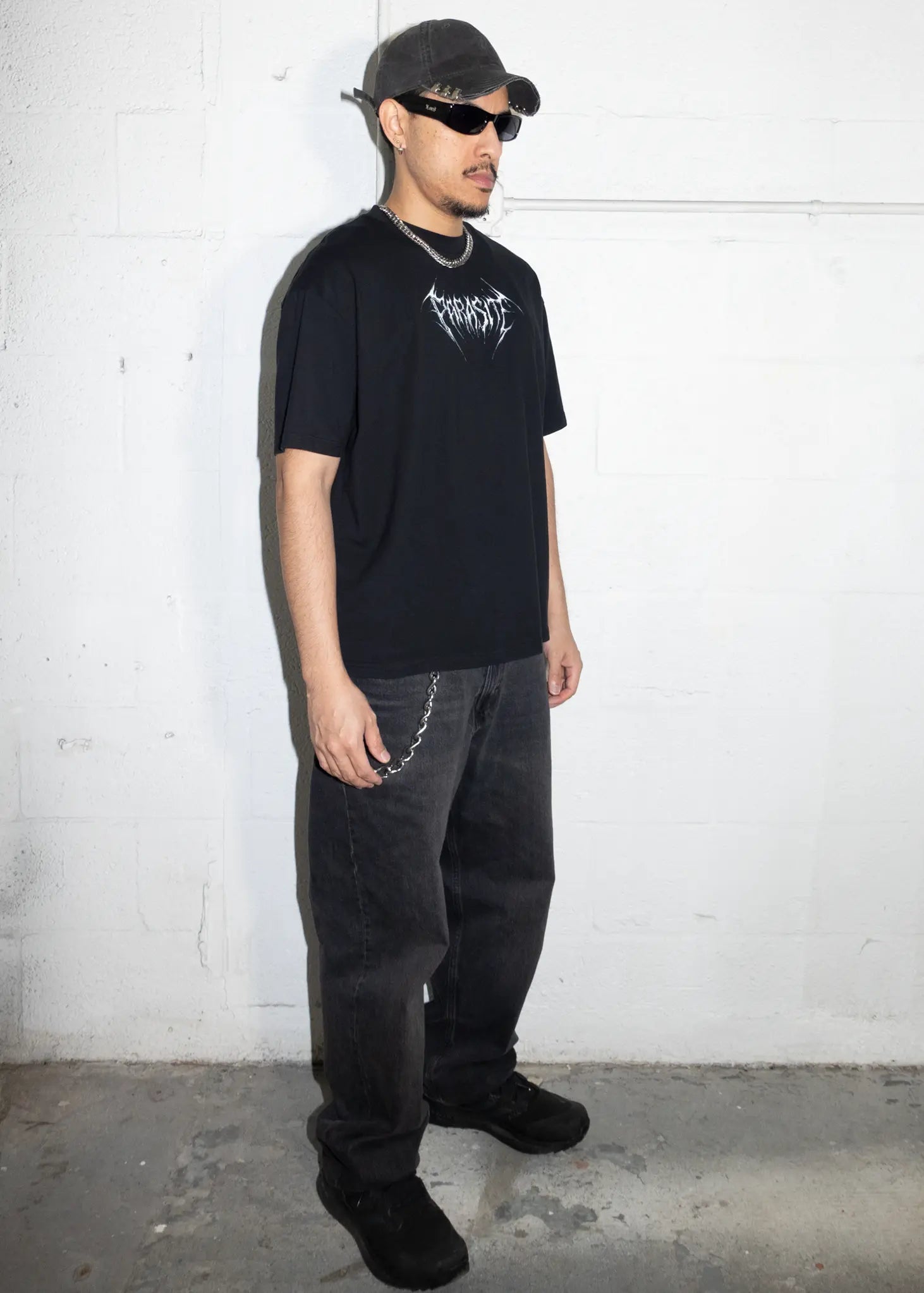 Person wearing a black t-shirt with text and dark pants against a white wall.