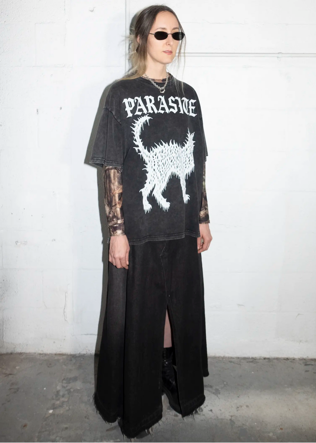 Person wearing a black Parasite Label text and Slime Cat t-shirt with a white graphic design against a white wall.