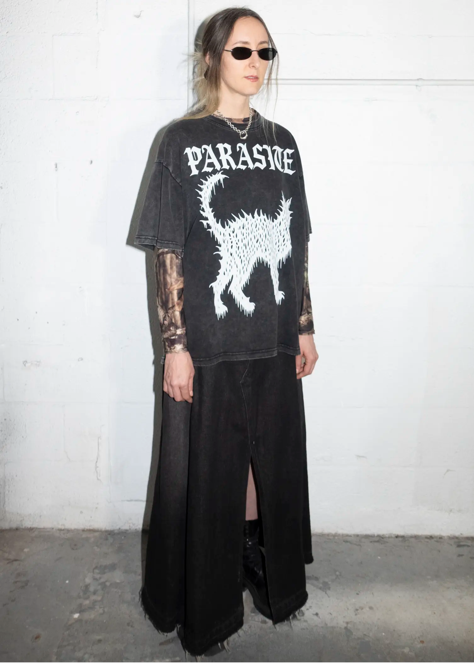 Person wearing a black Parasite Label text and Slime Cat t-shirt with a white graphic design against a white wall.