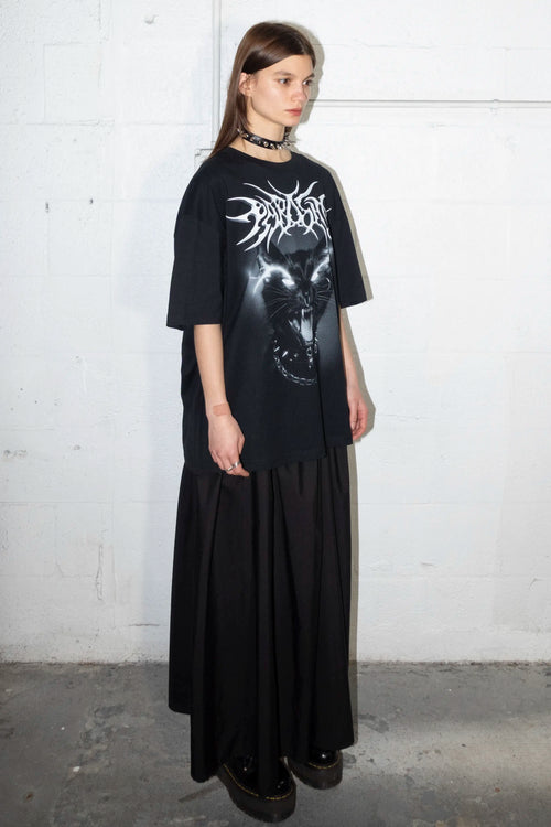Person wearing a black t-shirt with Parasite Label screaming punk cat graphic design design and long black skirt against a white wall.