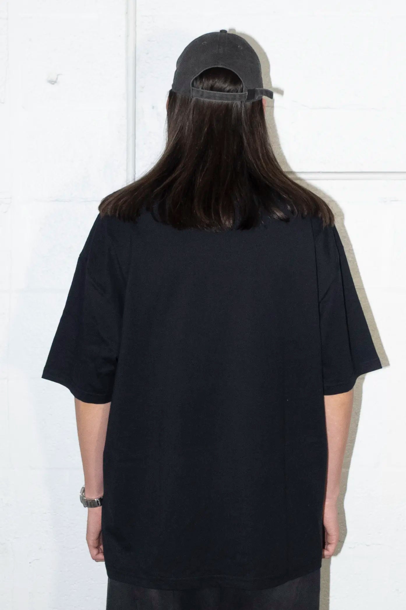Person wearing a black t-shirt and cap from behind against a white wall