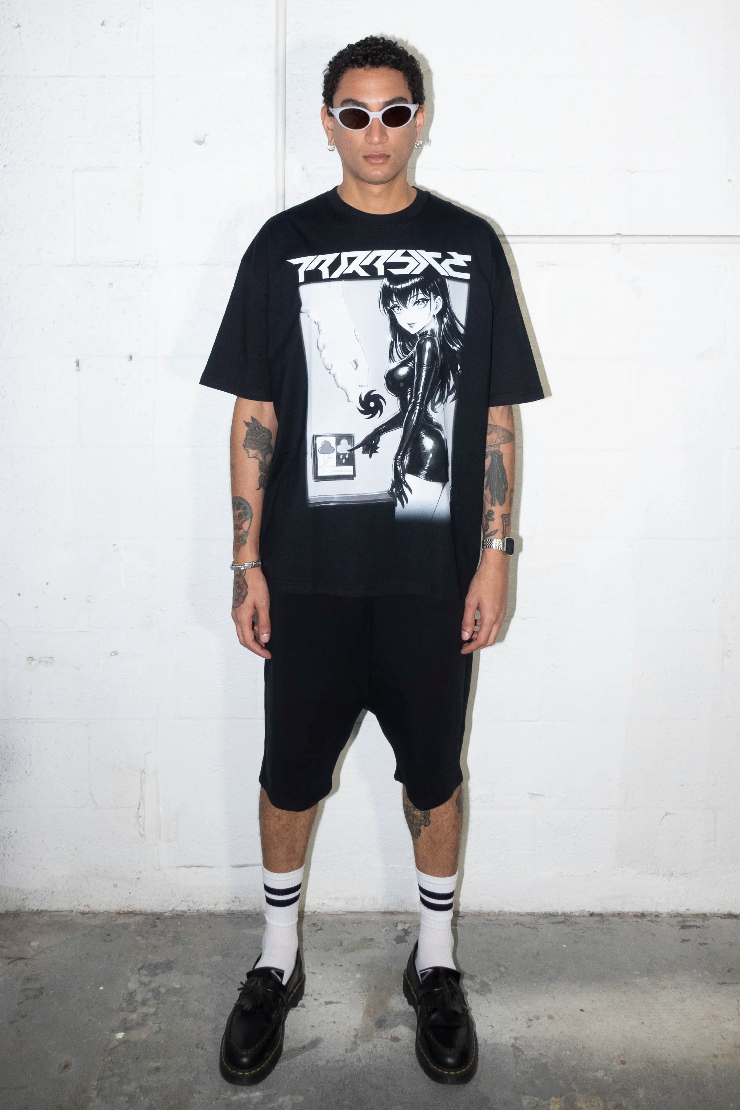 Person wearing a black t-shirt with a Parasite Label graphic design and text, standing against a white background black shorts, and black shoes against a white wall.