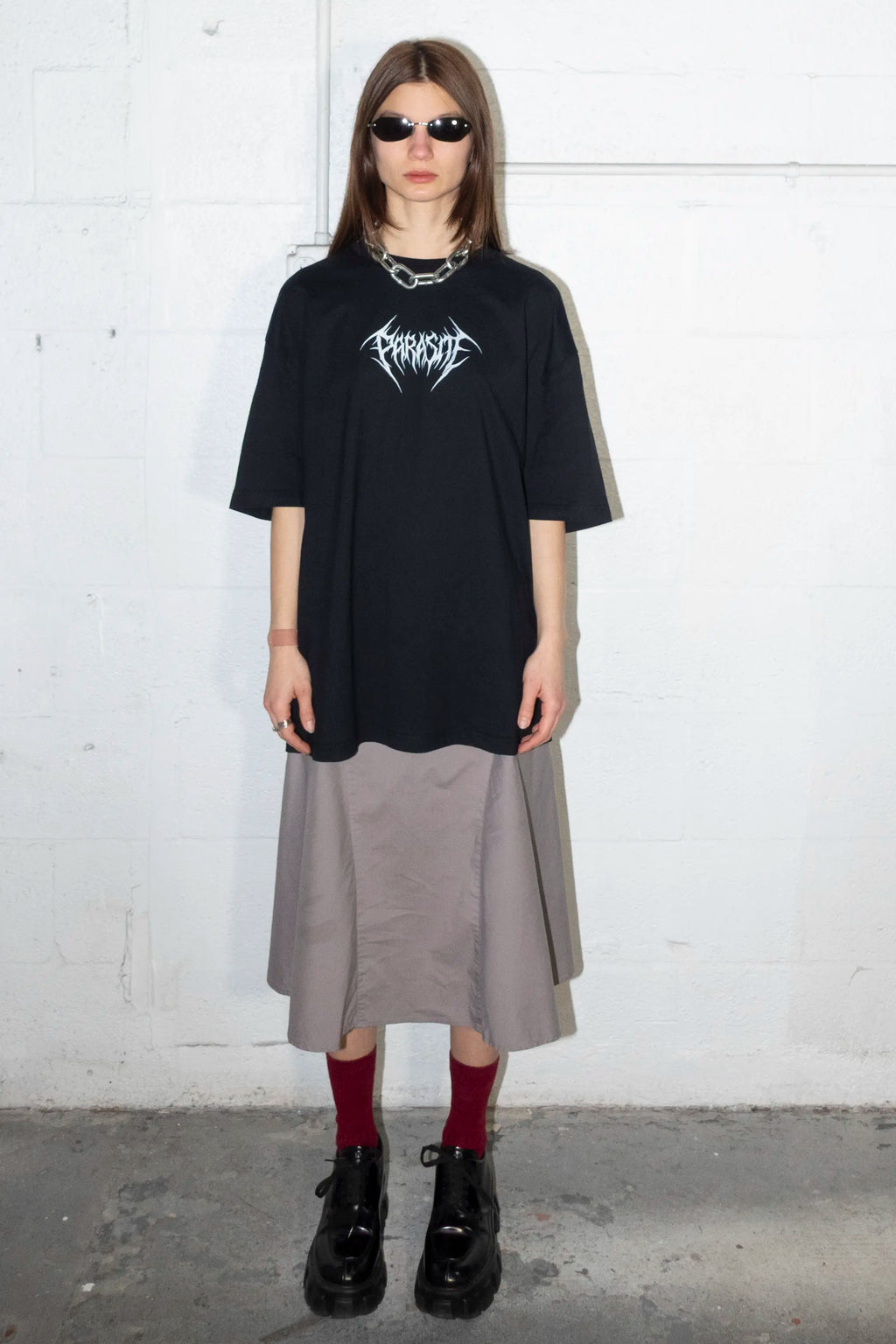 Person wearing a black oversized t-shirt with white Parasite Label text, gray skirt, red socks, and black shoes against a white wall.