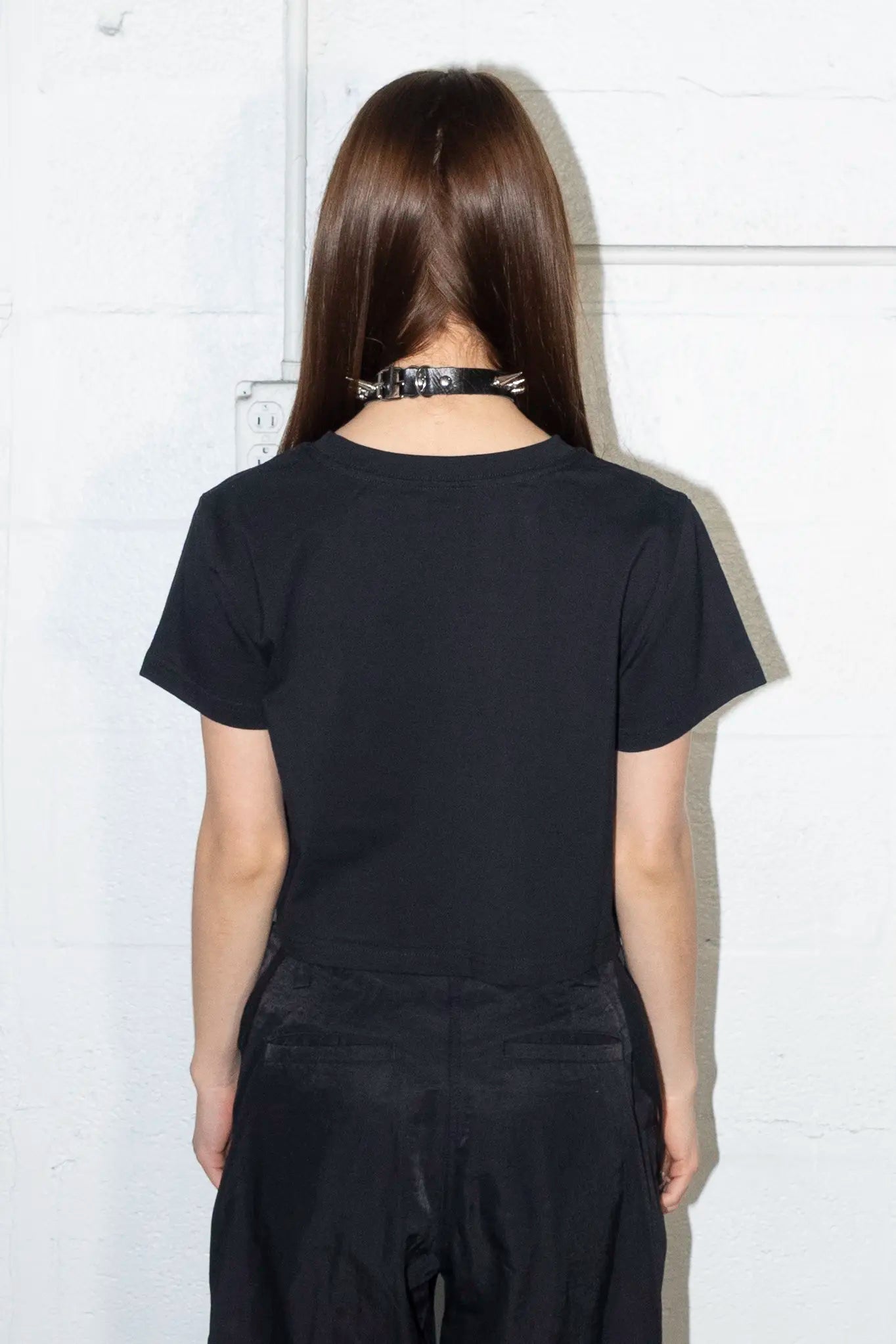 Person facing wall wearing a black t-shirt and pants with a choker, standing against a white wall. T-shirt no print on back.