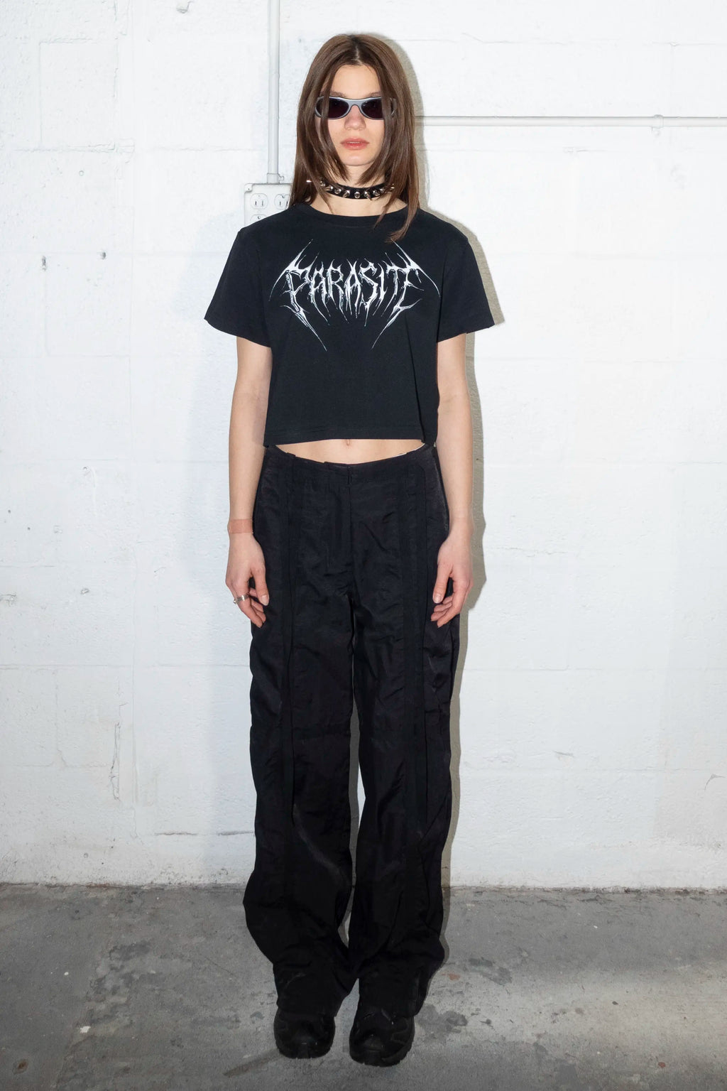 Person wearing a black t-shirt with a Parasite Label chrome 3d text logo graphic design and black pants against a white wall.