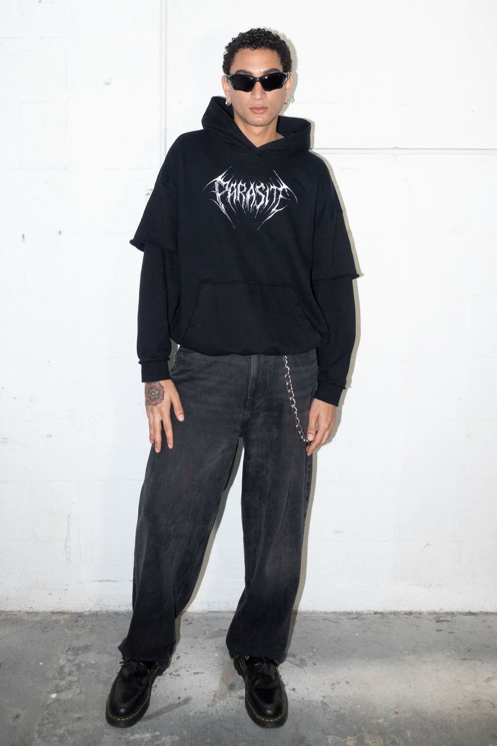 Person wearing a black hoodie with a 3d chrome Parasite Label logo and dark pants against a white wall.
