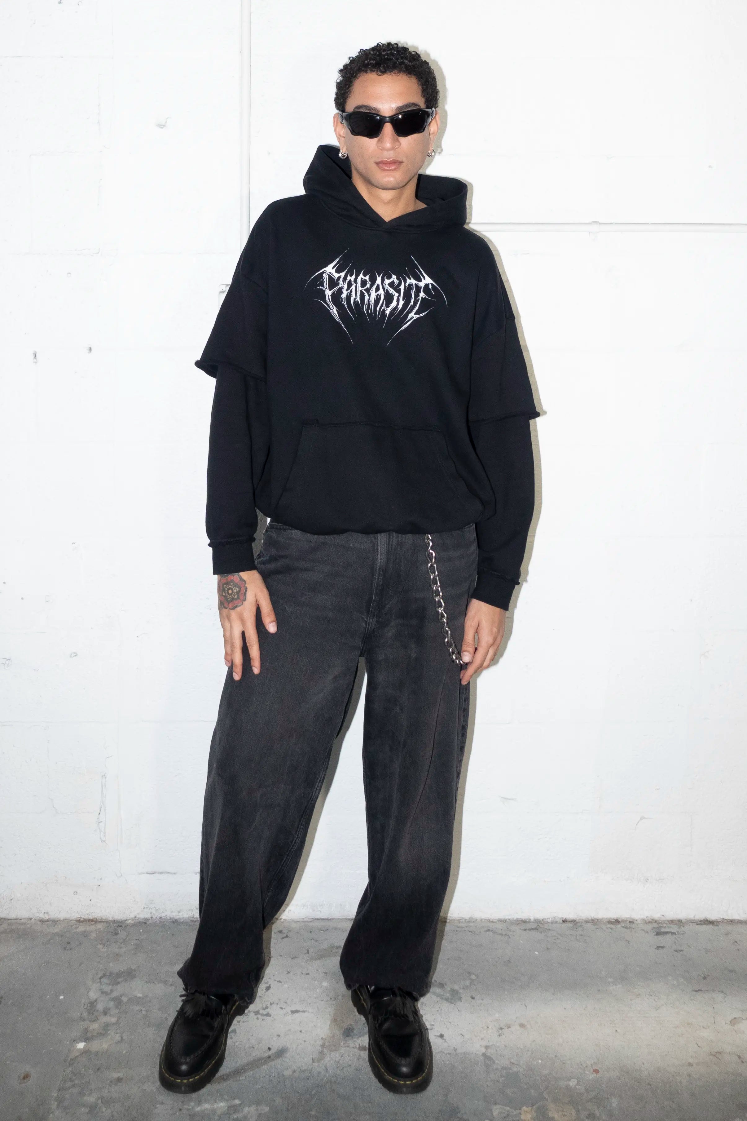 Person wearing a black hoodie with a 3d chrome Parasite Label logo and dark pants against a white wall.