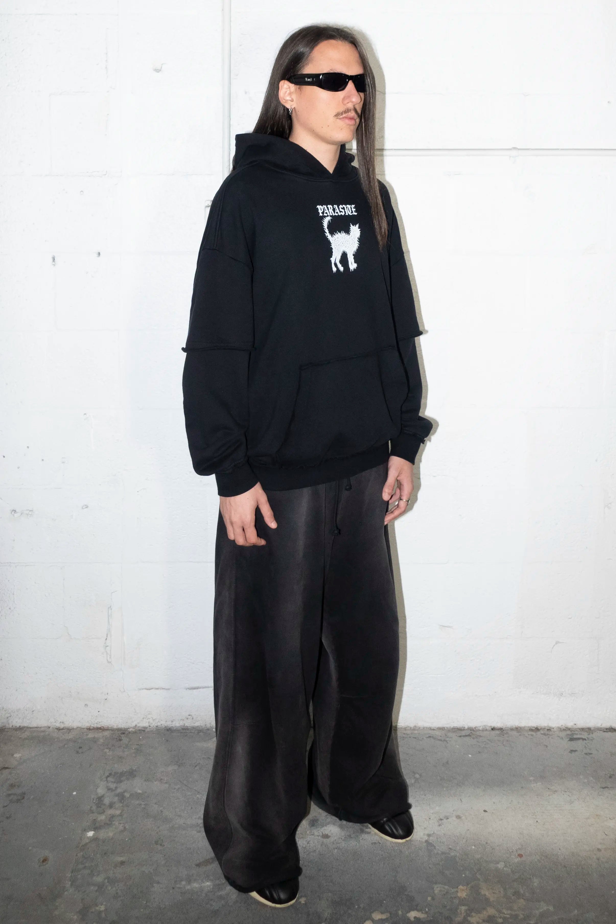 Person wearing a black hoodie with a Parasite Label logo and cat graphic design and black pants against a white wall.