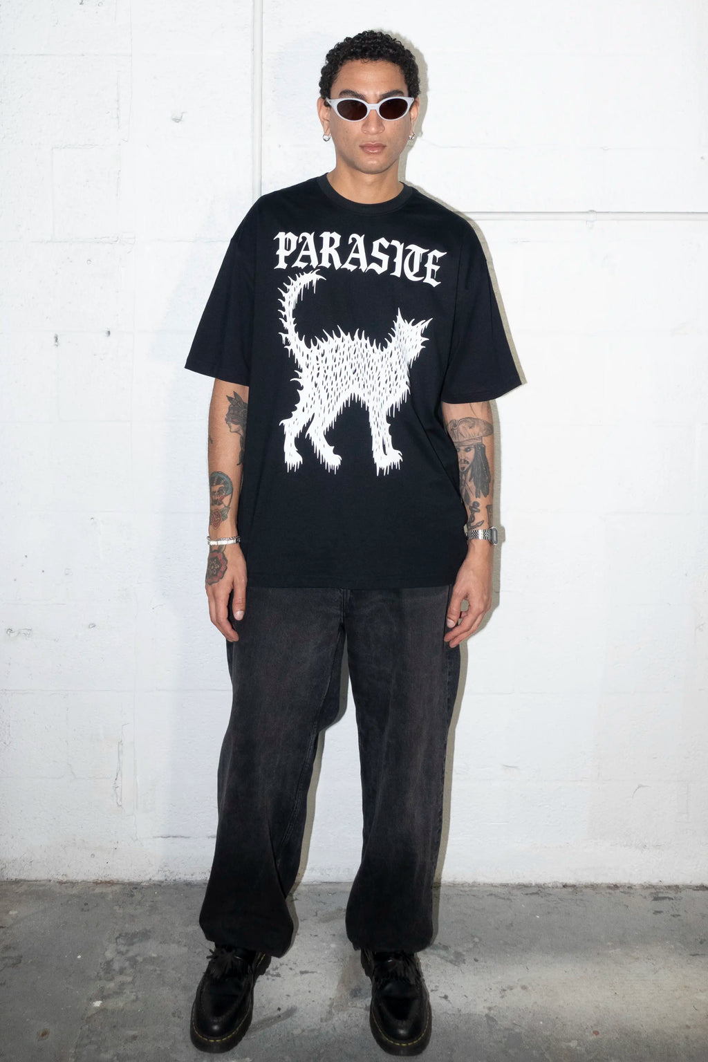 Person wearing a black t-shirt with a cat graphic design and Parasite Label text, standing against a white wall.