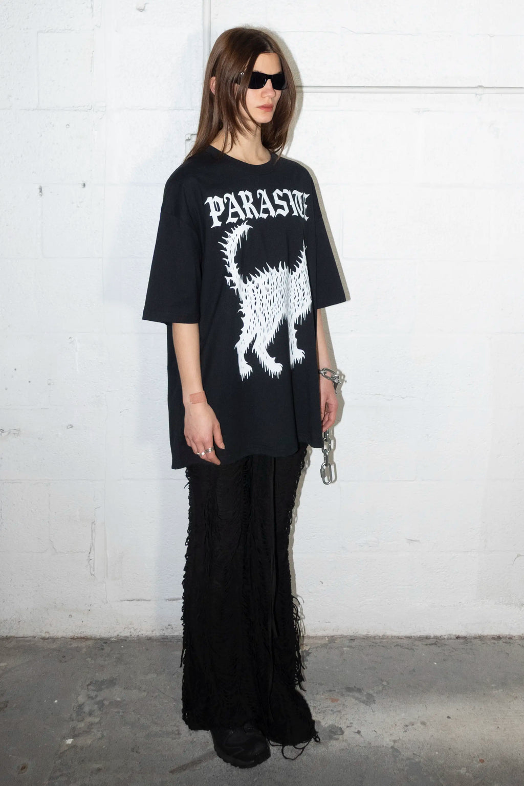 Person wearing a black oversized t-shirt with Parasite Label text logo and cat graphic design against a white wall.
