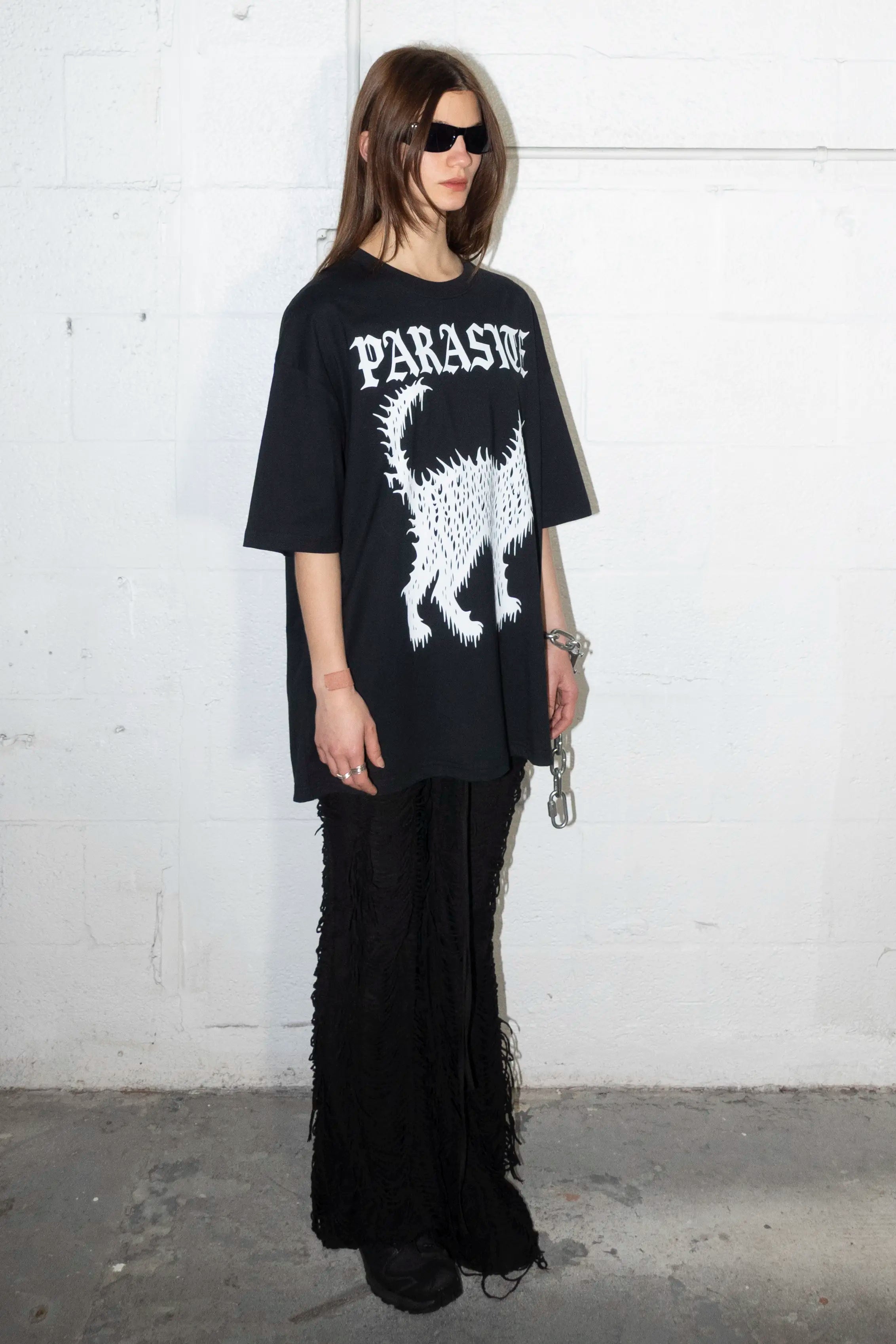 Person wearing a black oversized t-shirt with Parasite Label text logo and cat graphic design against a white wall.