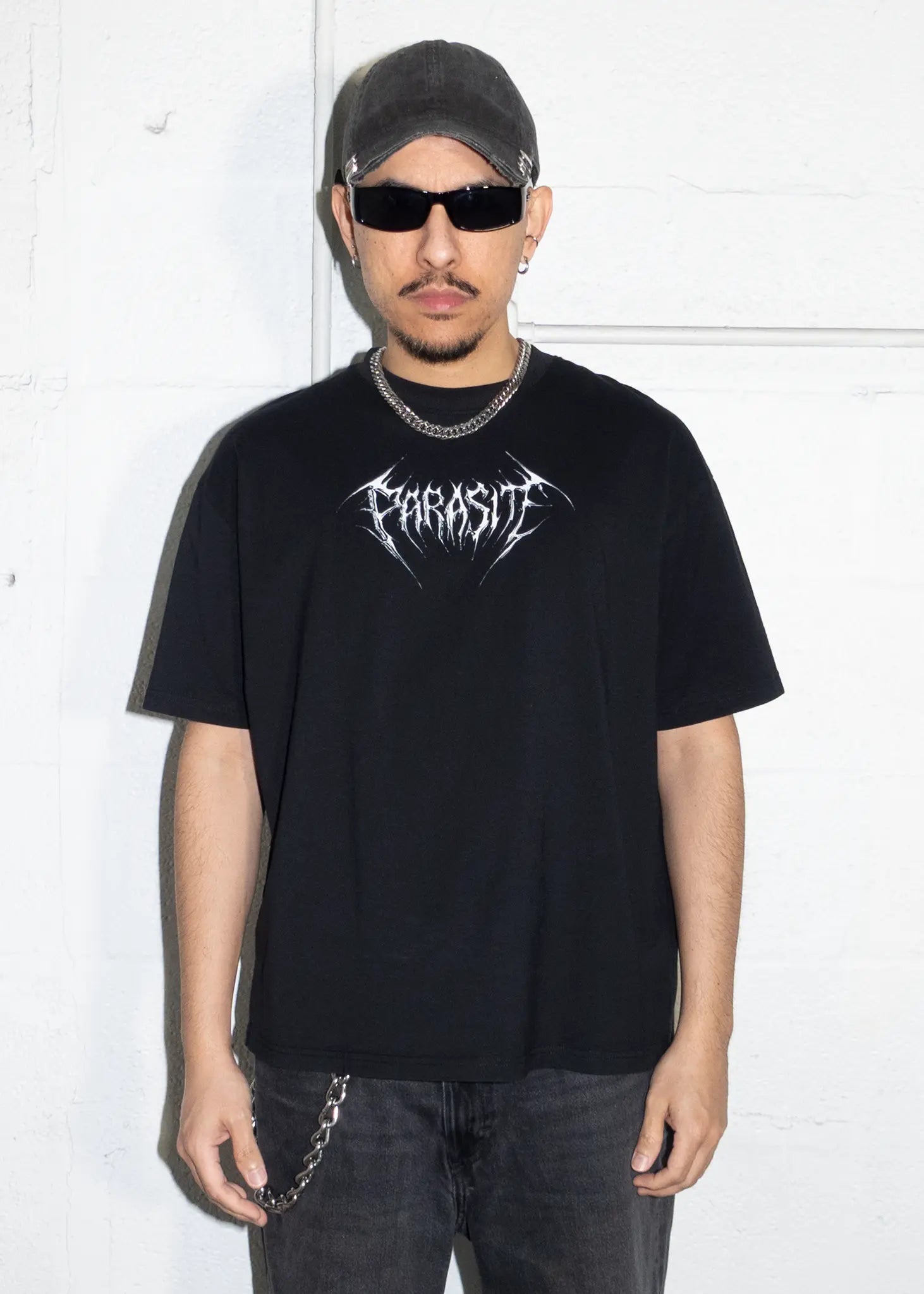 Person wearing a black t-shirt with a logo and dark sunglasses, standing against a white background.