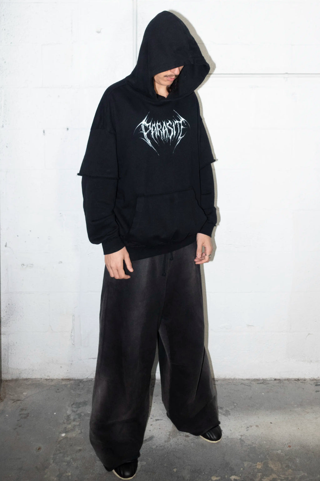 Person wearing a black hoodie with a 3d chrome Parasite Label logo and black pants against a white wall.
