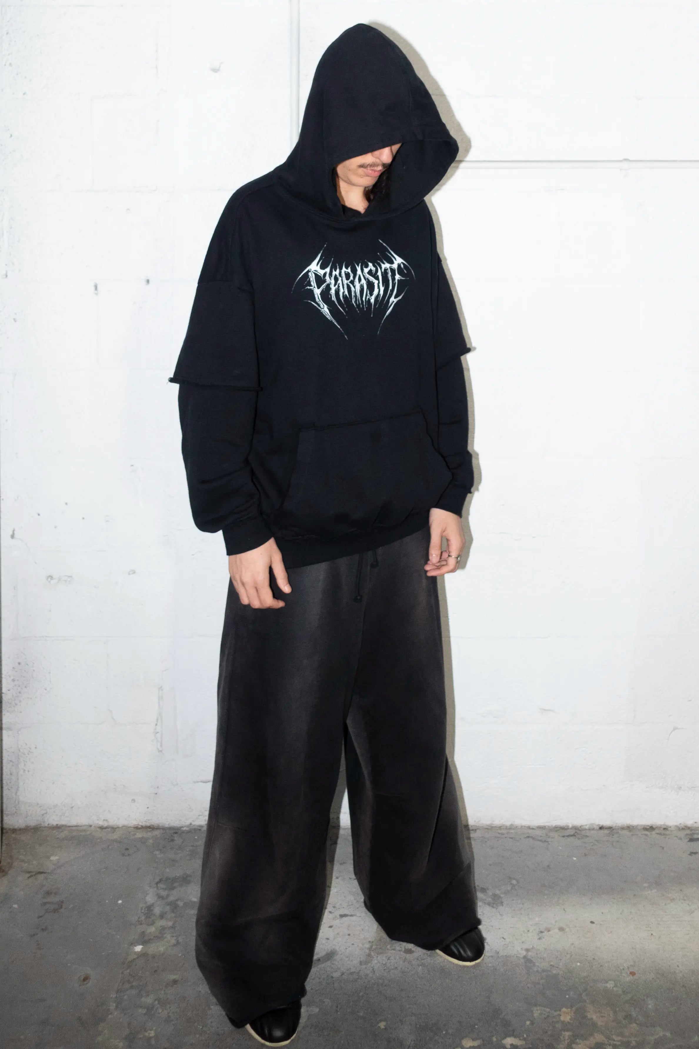 Person wearing a black hoodie with a 3d chrome Parasite Label logo and black pants against a white wall.