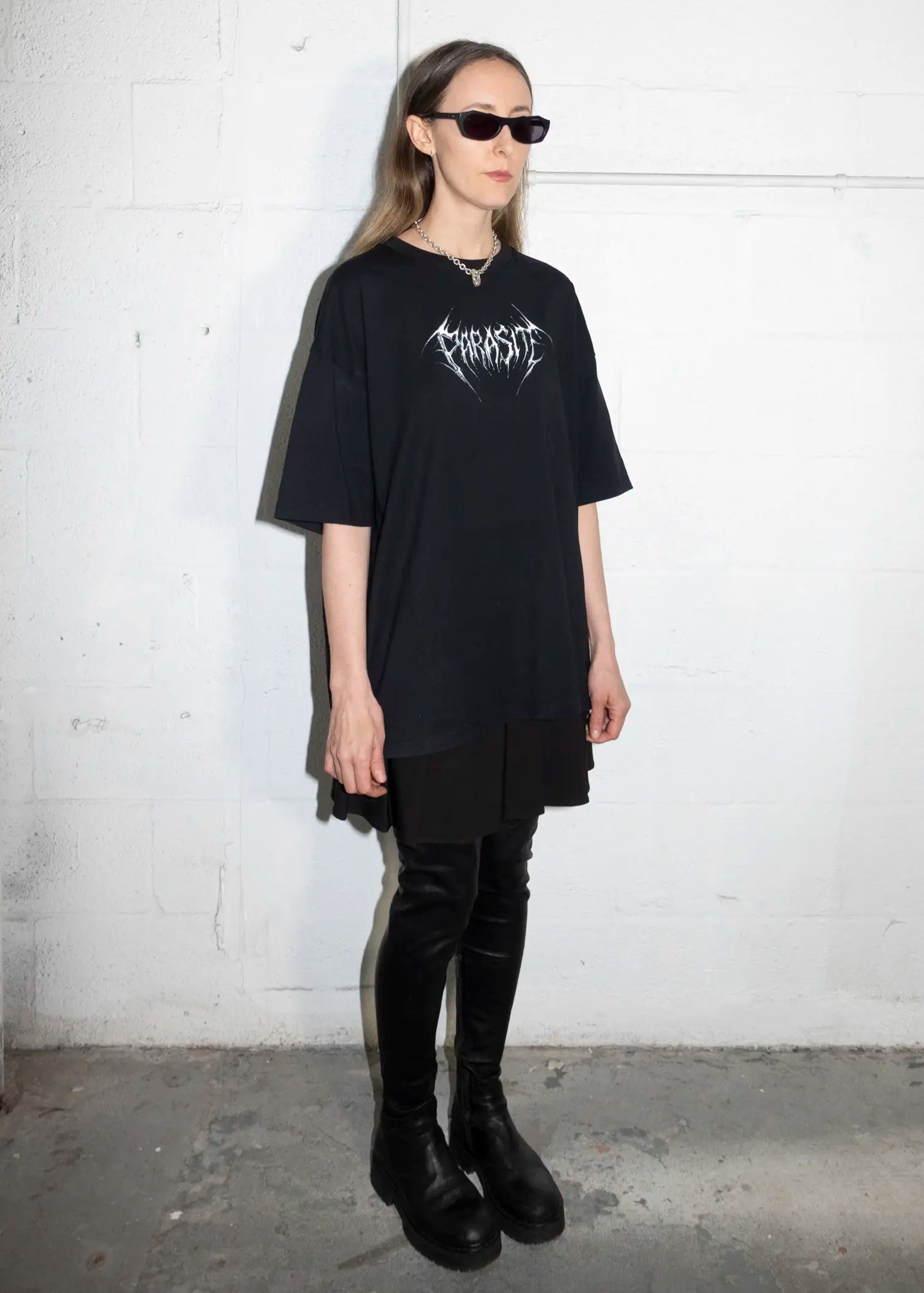 Person wearing a black oversized t-shirt with a logo, black pants, and black shoes against a white wall.