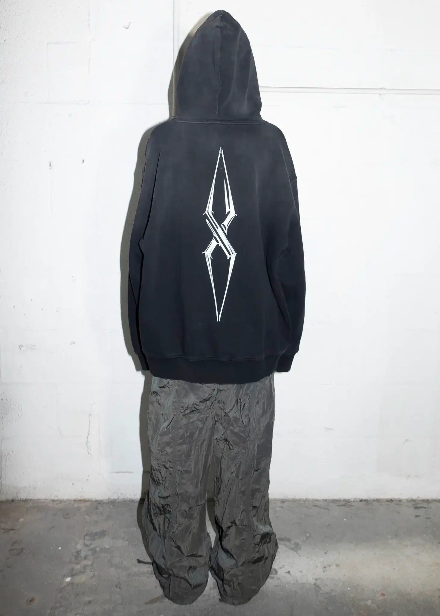 Person wearing a black hoodie with a white logo on the back, standing against a white wall.