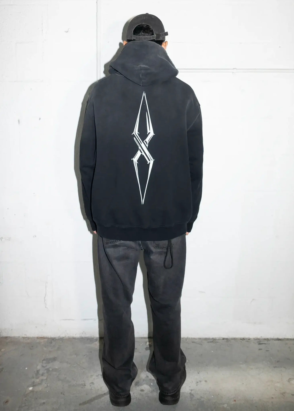 Person wearing a black hoodie with a white logo on the back against a white wall.