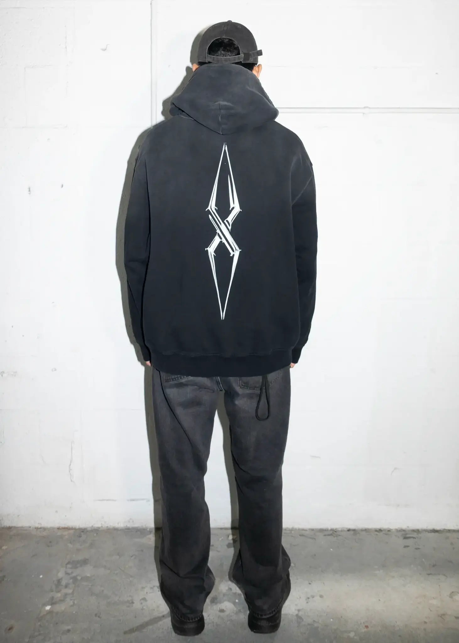 Person wearing a black hoodie with a white logo on the back against a white wall.