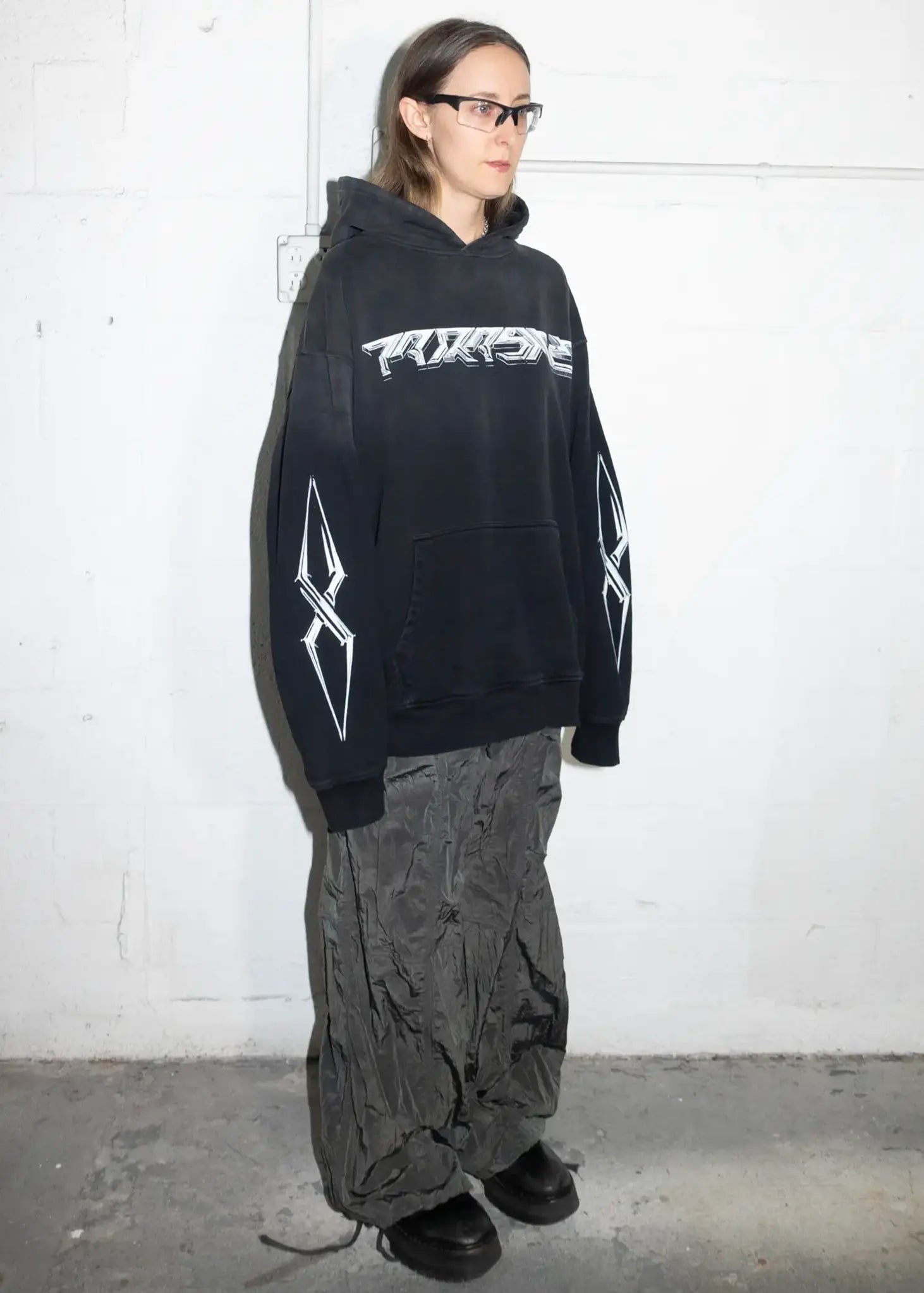 Person wearing a black hoodie with white text and graphics against a white wall.