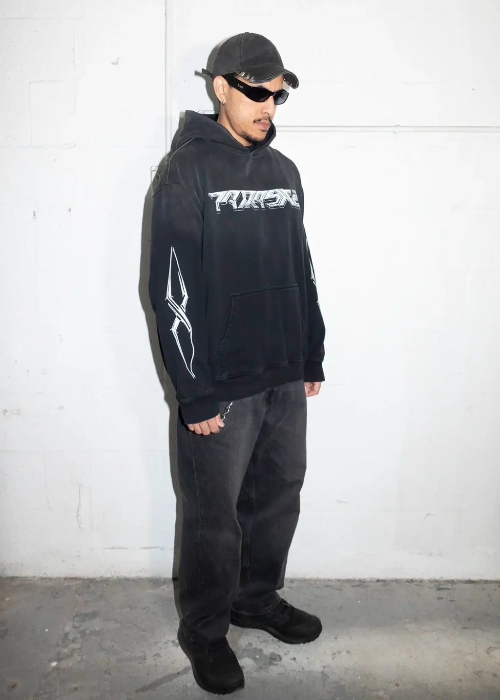 Person wearing a black hoodie with a logo, standing against a white wall.