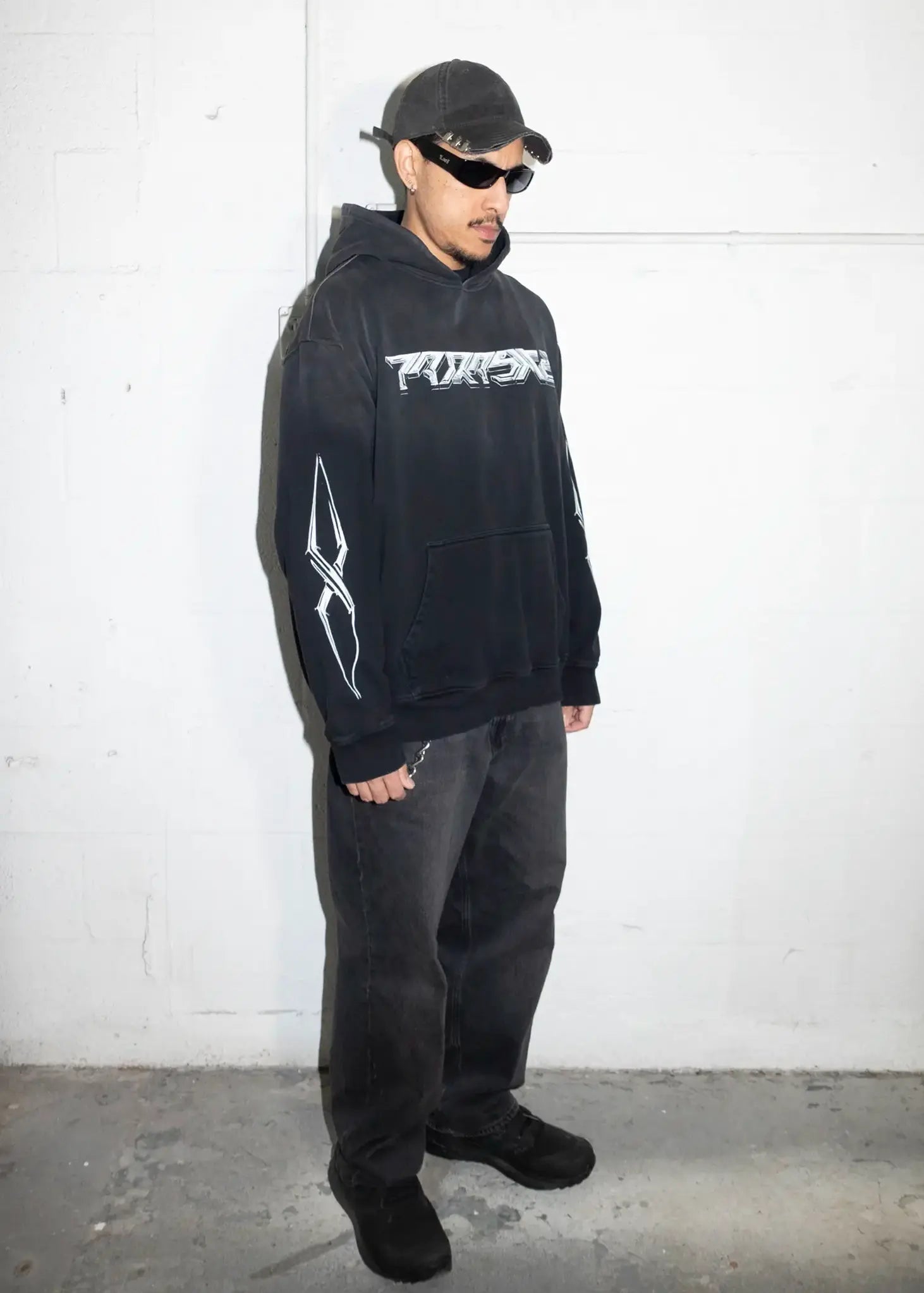 Person wearing a black hoodie with a logo, standing against a white wall.